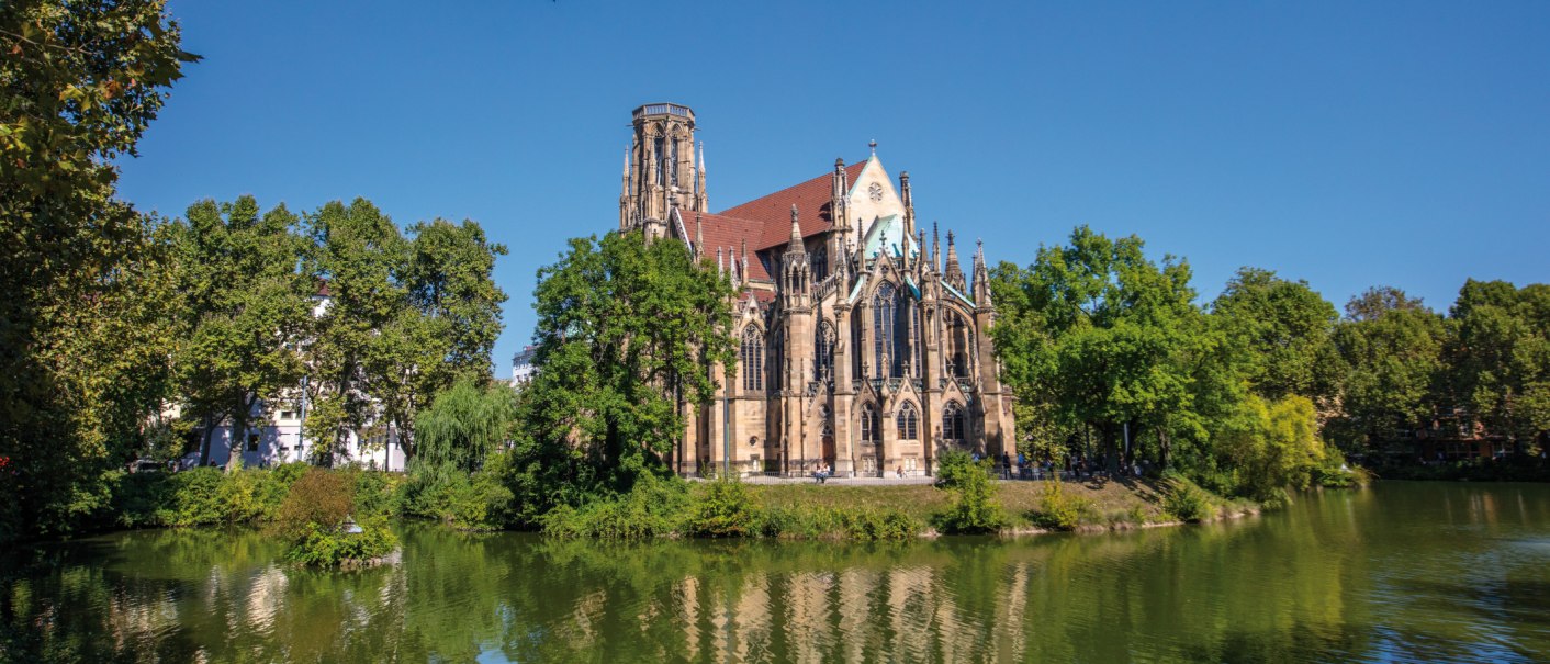 Die Johanneskirche am Feuersee in Stuttgart, umgeben von Bäumen und reflektiert im Wasser, bei klarem, sonnigem Wetter., © SMG Achim Mende Die Johanneskirche am Feuersee in Stuttgart, umgeben von Bäumen und reflektiert im Wasser, bei klarem, sonnigem Wetter., © SMG Achim Mende
