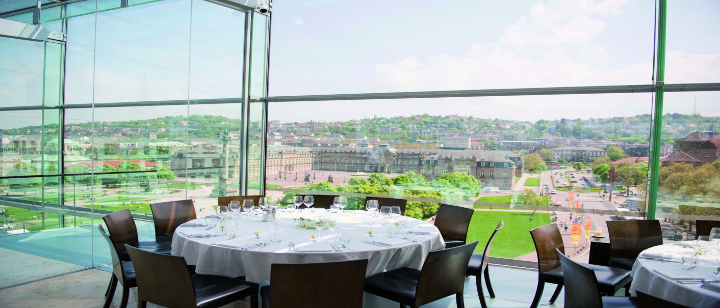 A round table with a white tablecloth and glasses stands in a glassed-in room with a view of a city and historic buildings., © Stuttgart-Marketing GmbH