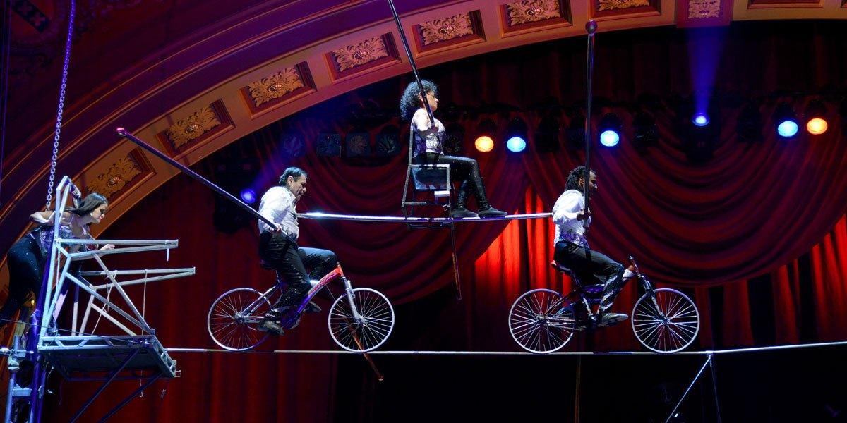Artisten balancieren auf Fahrrädern und einem Stuhl auf einem Hochseil im Zirkus, umgeben von rotem Vorhang und bunten Lichtern., © Thierry Bissat