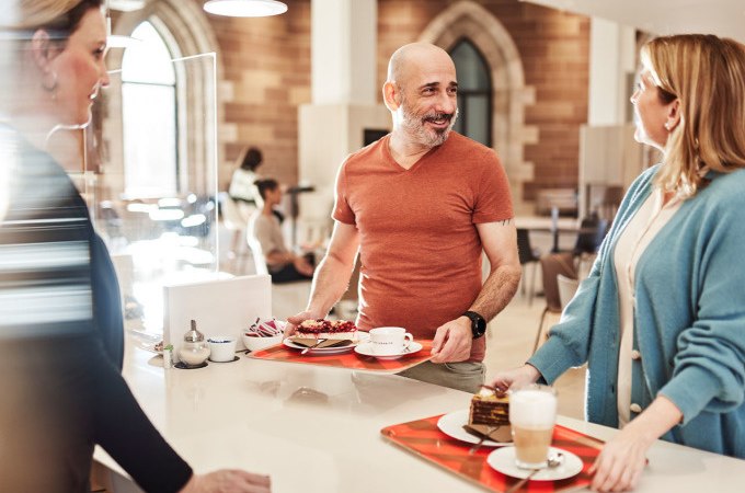 Drei Personen stehen in einem Café mit Tabletts voller Kaffee und Kuchen und unterhalten sich. Im Hintergrund sind große Fenster zu sehen., © Landesmuseum Württemberg Drei Personen stehen in einem Café mit Tabletts voller Kaffee und Kuchen und unterhalten sich. Im Hintergrund sind große Fenster zu sehen., © Landesmuseum Württemberg