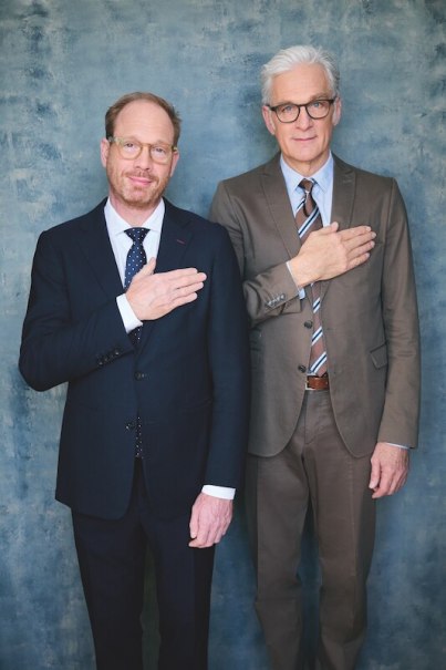 Two men in suits stand in front of a blue background, each holding a hand on their chest. They are wearing glasses and smiling slightly., &copy; Theaterhaus Stuttgart e.V.