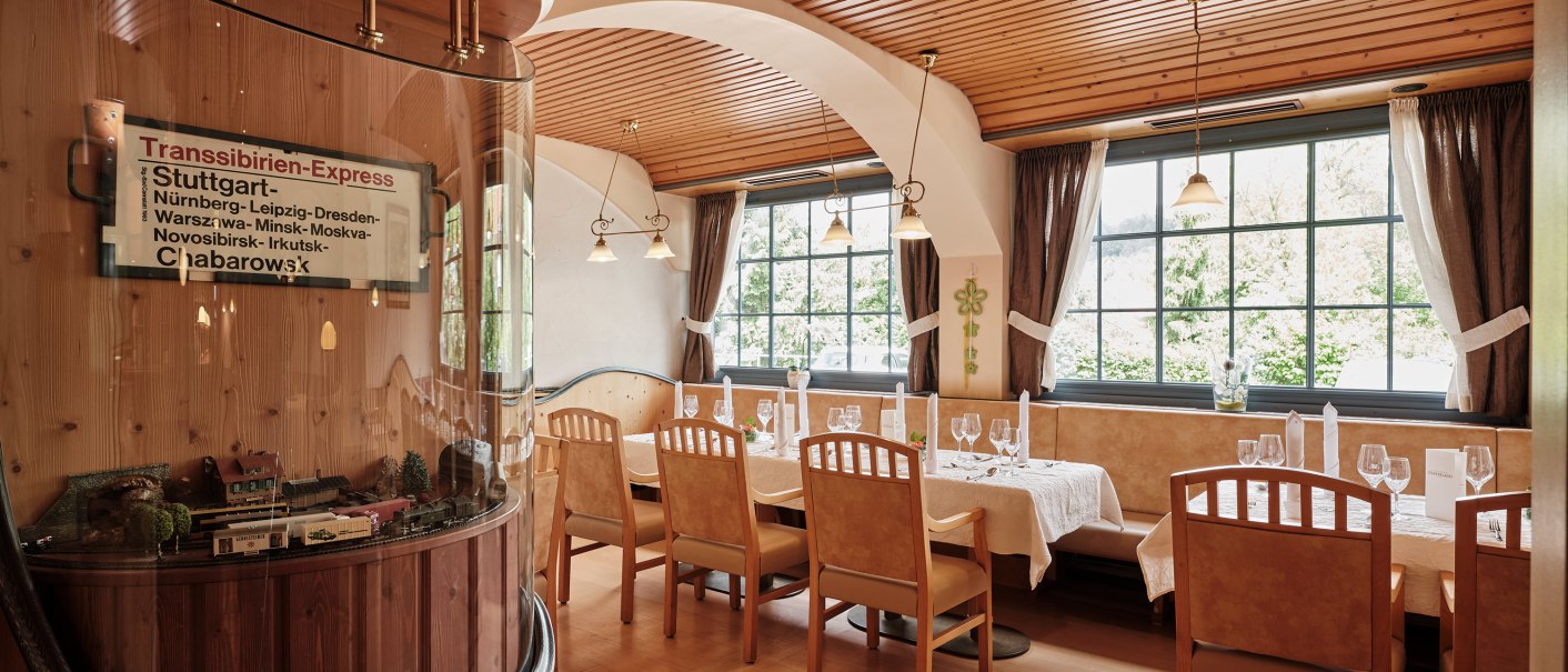 A cozy restaurant with wooden furniture, large windows, and a model train in a display case. Elegant table settings with glasses and napkins., © Badhotel Restaurant Stauferland A cozy restaurant with wooden furniture, large windows, and a model train in a display case. Elegant table settings with glasses and napkins., © Badhotel Restaurant Stauferland