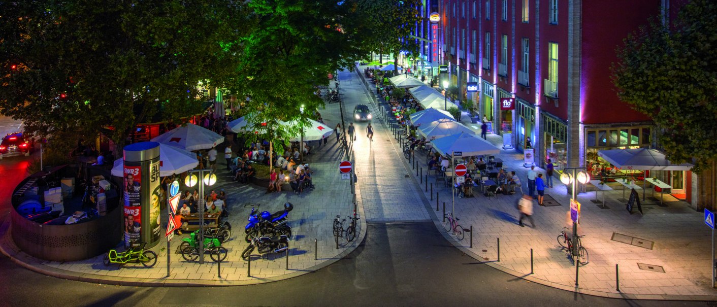 Belebte Stra&szlig;e bei Nacht mit Caf&eacute;s und Menschen. B&auml;ume und bunte Lichter schaffen eine lebendige Atmosph&auml;re. Fahrr&auml;der und Autos sind zu sehen., &copy; Stuttgart-Marketing GmbH