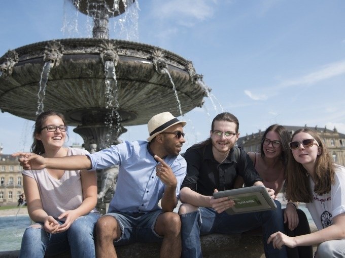 F&uuml;nf junge Erwachsene sitzen lachend vor einem Brunnen. Einer zeigt nach links, w&auml;hrend ein anderer ein Tablet h&auml;lt. Der Himmel ist klar und sonnig., &copy; ExitGames Stuttgart