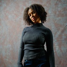 Eine Frau mit lockigem Haar trägt einen grauen Pullover und steht vor einem neutralen Hintergrund. Sie blickt leicht zur Seite., © links im Bild Eine Frau mit lockigem Haar trägt einen grauen Pullover und steht vor einem neutralen Hintergrund. Sie blickt leicht zur Seite., © links im Bild