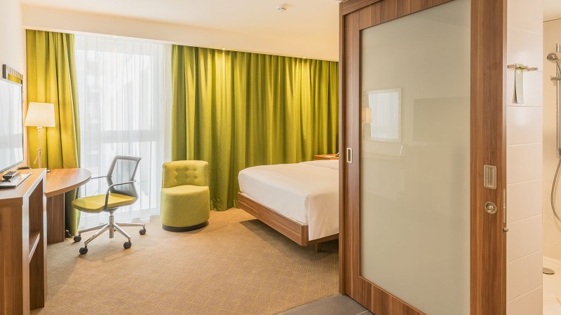 A modern hotel room with a double bed, a green armchair, a desk, and a view into the bathroom with a shower. Light-colored curtains and green drapes., © Hampton by Hilton Stuttgart City Centre A modern hotel room with a double bed, a green armchair, a desk, and a view into the bathroom with a shower. Light-colored curtains and green drapes., © Hampton by Hilton Stuttgart City Centre