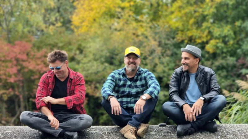 Drei Männer sitzen entspannt auf einer Mauer im Freien. Sie tragen lässige Kleidung und sind von herbstlichen Bäumen umgeben., © Laszlo Váray Drei Männer sitzen entspannt auf einer Mauer im Freien. Sie tragen lässige Kleidung und sind von herbstlichen Bäumen umgeben., © Laszlo Váray