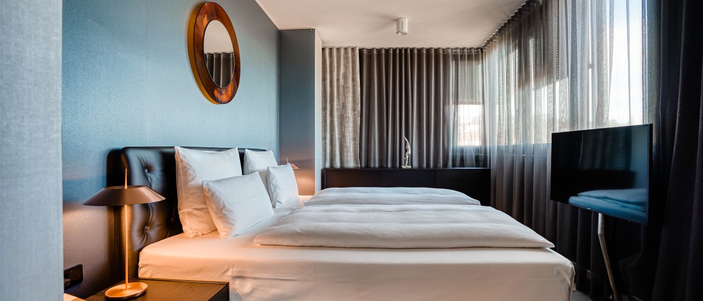 Modernes Hotelzimmer mit Doppelbett, Spiegel an der Wand, Nachttischlampen und Fernseher. Große Fenster mit Vorhängen., © CLOUD N°7 APARTMENTS GmbH Modernes Hotelzimmer mit Doppelbett, Spiegel an der Wand, Nachttischlampen und Fernseher. Große Fenster mit Vorhängen., © CLOUD N°7 APARTMENTS GmbH