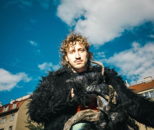 Eine Person in einem schwarzen, fellartigen Kost&uuml;m mit H&ouml;rnern steht vor einem blauen Himmel mit Wolken. Im Hintergrund sind Geb&auml;ude zu sehen., &copy; Rosenau Kultur e.V.