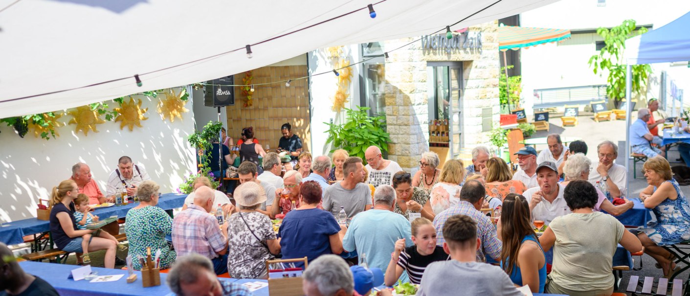 A social gathering outdoors at the Zaiß Winery. People are enjoying food and drinks under a sunshade., © Weingut Zaiß A social gathering outdoors at the Zaiß Winery. People are enjoying food and drinks under a sunshade., © Weingut Zaiß