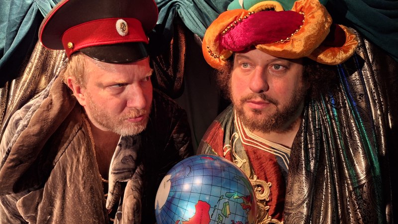 Zwei M&auml;nner in fantasievollen Kost&uuml;men, einer mit roter M&uuml;tze, der andere mit buntem Turban, halten einen Globus. Der Hintergrund ist mit Stoff drapiert., &copy; Theater am Olgaeck