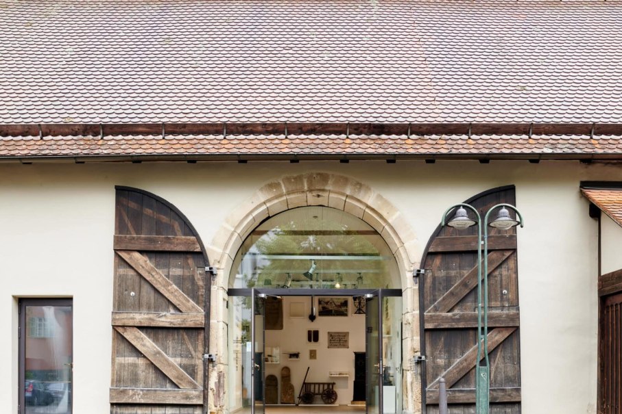 Eingang des Heimatmuseums Plieningen mit großen, offenen Holztüren und einer Glasfront. Innen sind Ausstellungsstücke sichtbar., © StadtPalais / svenweber_repro_ret-min Eingang des Heimatmuseums Plieningen mit großen, offenen Holztüren und einer Glasfront. Innen sind Ausstellungsstücke sichtbar., © StadtPalais / svenweber_repro_ret-min