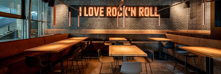 A stylish café with wooden tables and modern chairs. The words 'I Love Rock 'n Roll' light up the wall in large letters., © Maks Richter