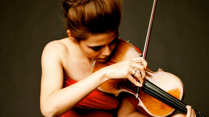 Eine Frau in einem roten Kleid spielt konzentriert Geige. Der Hintergrund ist dunkel, was den Fokus auf die Musikerin und ihr Instrument lenkt., © Gabrielle Revere Eine Frau in einem roten Kleid spielt konzentriert Geige. Der Hintergrund ist dunkel, was den Fokus auf die Musikerin und ihr Instrument lenkt., © Gabrielle Revere