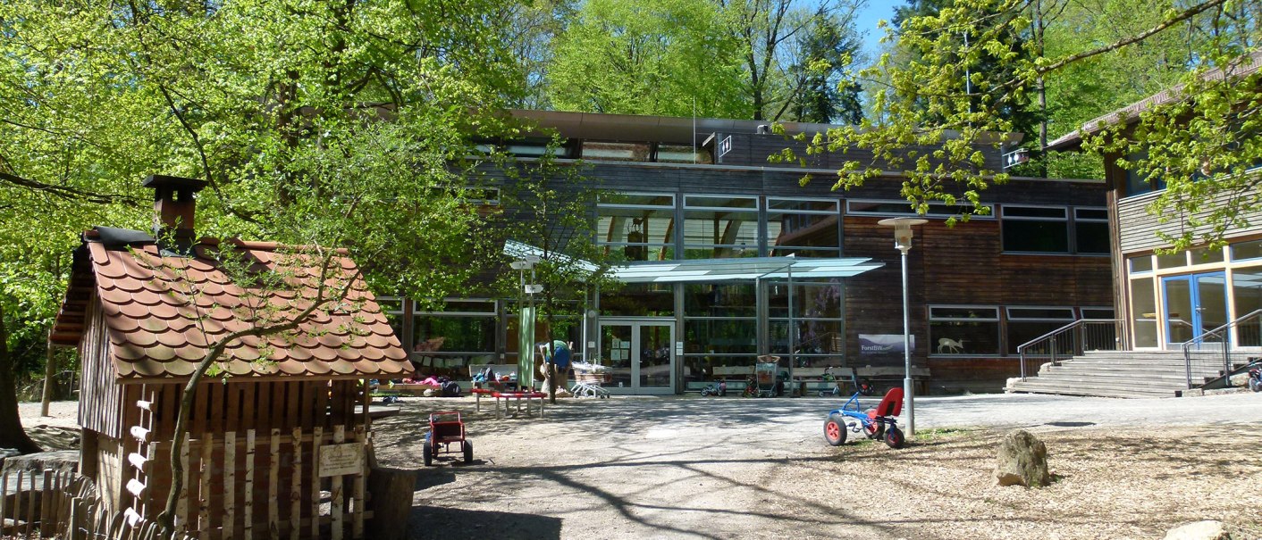 Inner courtyard of the House of the Forest with toy vehicles, trees and a small wooden house. Modern glass fa&ccedil;ade in the background., &copy; Haus des Waldes