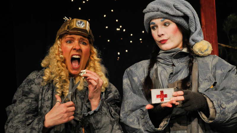 Zwei Schauspielerinnen in grauen Kostümen auf der Bühne. Eine hält ein Erste-Hilfe-Set, die andere zeigt einen schmerzhaften Gesichtsausdruck., © TheaterFusion