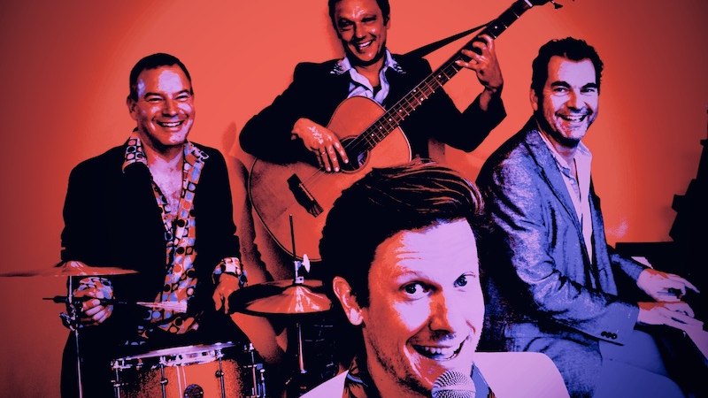 Vier Männer in Anzügen musizieren fröhlich. Einer singt ins Mikrofon, die anderen spielen Schlagzeug, Gitarre und Klavier. Der Hintergrund ist orange., © Rosenau Kultur e.V. Vier Männer in Anzügen musizieren fröhlich. Einer singt ins Mikrofon, die anderen spielen Schlagzeug, Gitarre und Klavier. Der Hintergrund ist orange., © Rosenau Kultur e.V.