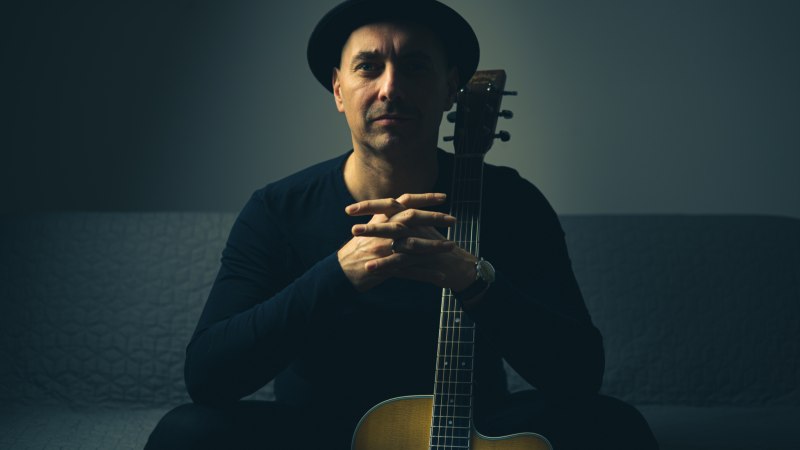 Ein Mann mit schwarzem Hut sitzt auf einem Sofa und hält eine Akustikgitarre. Die Beleuchtung ist gedämpft und erzeugt eine ruhige Atmosphäre., © Foto: Tom Lumen Ein Mann mit schwarzem Hut sitzt auf einem Sofa und hält eine Akustikgitarre. Die Beleuchtung ist gedämpft und erzeugt eine ruhige Atmosphäre., © Foto: Tom Lumen