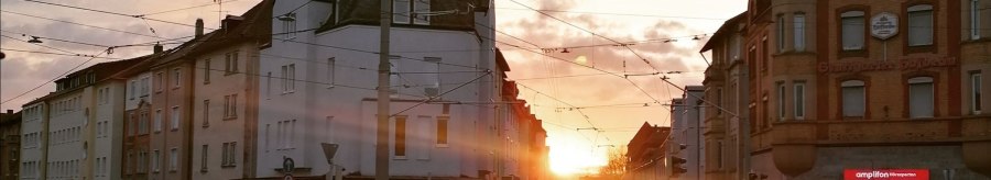 Sonnenuntergang in einer Stadtstra&szlig;e mit historischen Geb&auml;uden und Stra&szlig;enbahnkabeln. Menschen &uuml;berqueren die Stra&szlig;e, w&auml;hrend die Sonne zwischen den H&auml;usern untergeht., &copy; SMG