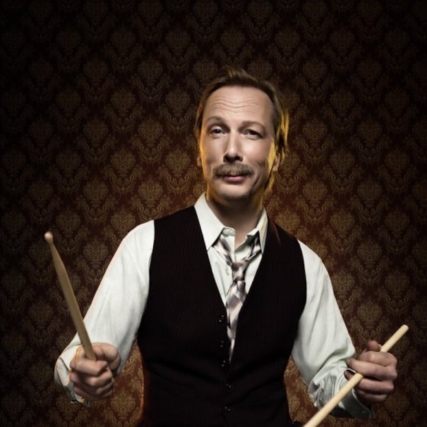 A man with a moustache holds drumsticks in front of a patterned wallpaper. He is wearing a vest and a shirt with a tie., &copy; Music Circus Concertb&uuml;ro GmbH &CoKG