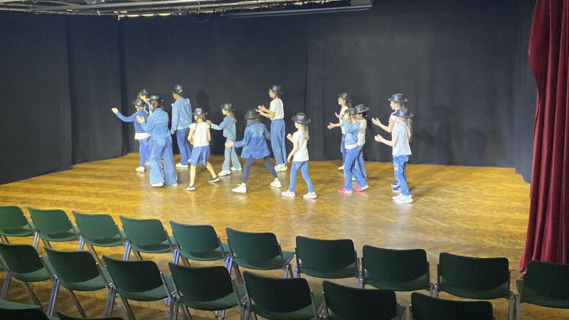 Eine Gruppe von Kindern tanzt auf einer Bühne in einem leeren Theater. Sie tragen Hüte und lässige Kleidung. Der Raum ist mit grünen Stühlen bestuhlt., © Tathea - Lena Oschowitzer Eine Gruppe von Kindern tanzt auf einer Bühne in einem leeren Theater. Sie tragen Hüte und lässige Kleidung. Der Raum ist mit grünen Stühlen bestuhlt., © Tathea - Lena Oschowitzer