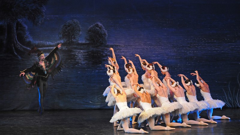 Ballettszene aus "Schwanensee" mit Tänzern in weißen Tutus und einem Tänzer in schwarzem Kostüm vor einer See-Kulisse., © Go 2 - Convent GmbH Ballettszene aus "Schwanensee" mit Tänzern in weißen Tutus und einem Tänzer in schwarzem Kostüm vor einer See-Kulisse., © Go 2 - Convent GmbH