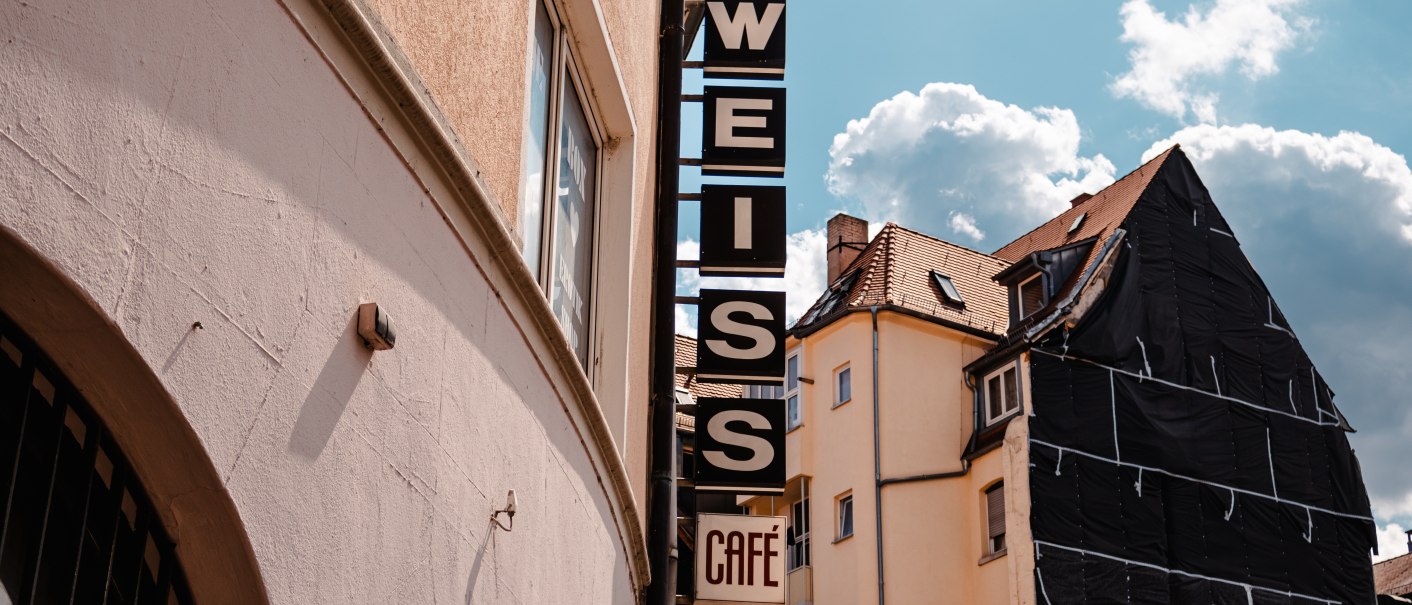 Schild mit 'WEISS Café' an einem Gebäude, daneben ein weiteres Haus mit Gerüst. Blauer Himmel mit Wolken im Hintergrund., © SMG_Sarah Schmid