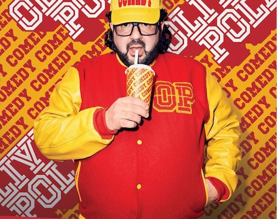 Ein Mann in roter und gelber Jacke mit einer 'Comedy'-Mütze trinkt aus einem Becher. Der Hintergrund zeigt den Namen 'Oliver Polak'., © Theaterhaus Stuttgart e.V.