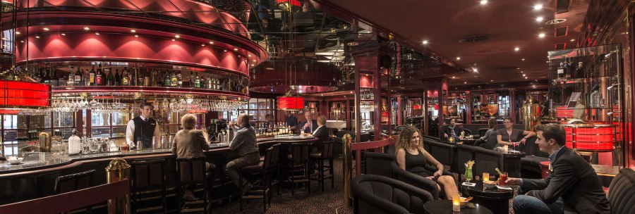 Elegante Bar mit rotem Dekor, Spiegeln und ged&auml;mpfter Beleuchtung. G&auml;ste sitzen an der Theke und an Tischen, w&auml;hrend ein Barkeeper Getr&auml;nke serviert., &copy; Maritim Hotels