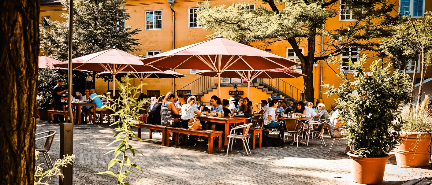 Menschen sitzen unter roten Sonnenschirmen in einem sonnigen Außenbereich vor einem gelben Gebäude., © Amadeus