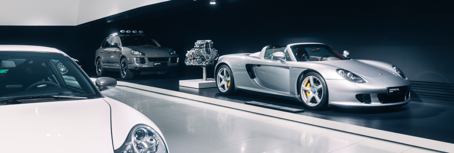 Three Porsche models in a modern showroom: a silver sports car, an SUV and a white car in the foreground. An engine is also on display., &copy; Porsche AG