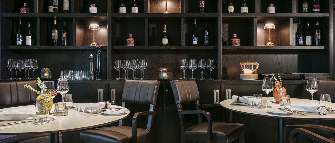 Elegantes Restaurant mit gedeckten Tischen, Weingläsern und einem Regal voller Weinflaschen und Dekorationen im Hintergrund., © Arcotel Hotels
