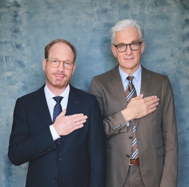 Two men in suits stand next to each other in front of a blue background, each holding a hand on their chest., &copy; Theaterhaus Stuttgart e.V.
