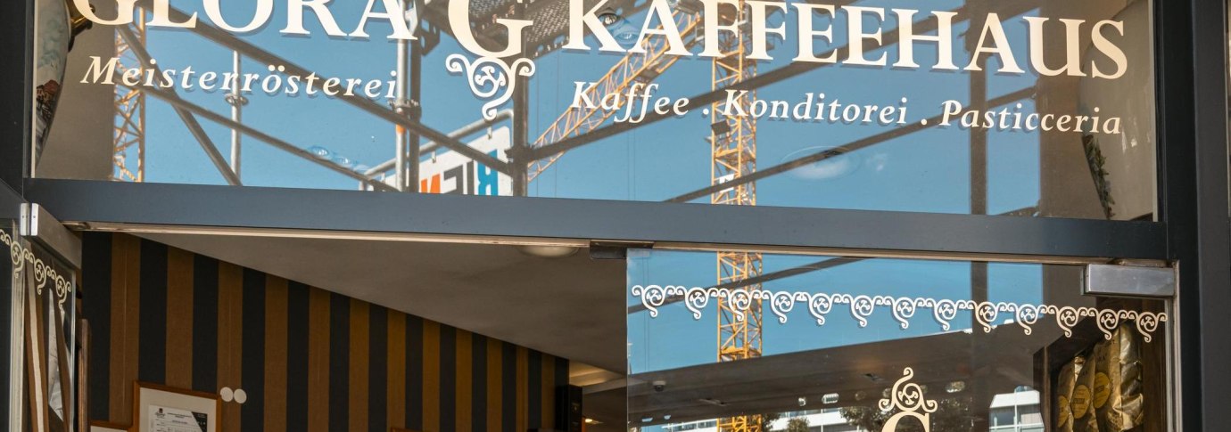 Eingang des Glora Kaffeehauses mit dekorativem Schriftzug auf der Glasfront. Im Hintergrund sind Baukr&auml;ne zu sehen., &copy; SMG, Sarah Schmid