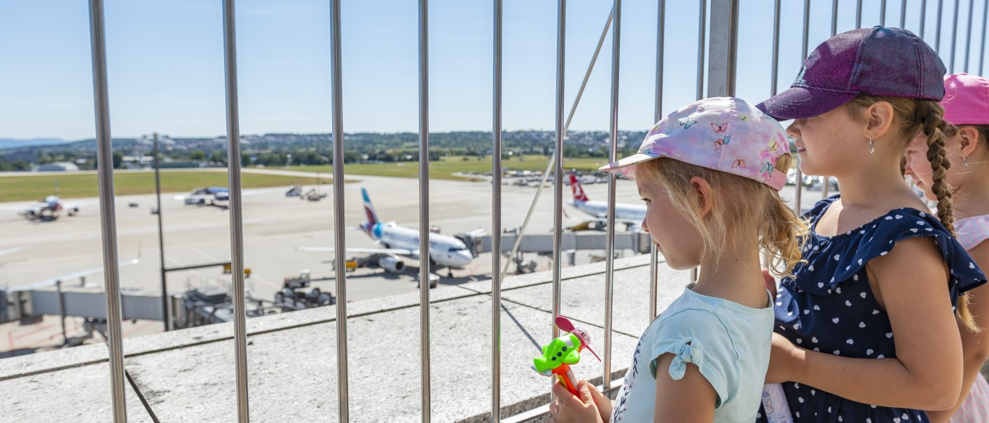 Kinder stehen auf der Besucherterrasse des Stuttgarter Flughafens und schauen durch ein Gitter auf das Rollfeld mit Flugzeugen., © Flughafen Stuttgart Kinder stehen auf der Besucherterrasse des Stuttgarter Flughafens und schauen durch ein Gitter auf das Rollfeld mit Flugzeugen., © Flughafen Stuttgart