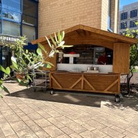 Ein Holzstand vor einem modernen Gebäude mit Glasfassade. Im Vordergrund sind Pflanzen zu sehen, die den Stand teilweise verdecken., © TOMAS