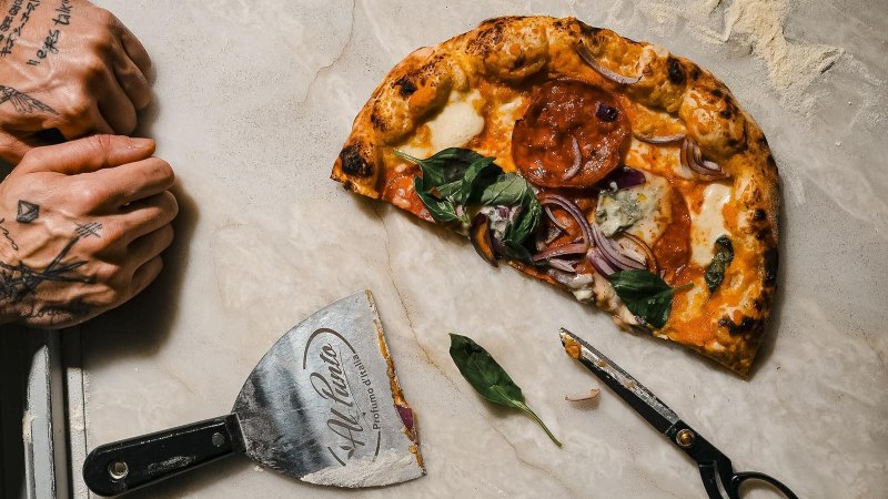 Halb gegessene Pizza mit Basilikum und Zwiebeln auf hellem Stein, daneben Pizzaschneider und Schere; links zwei t&auml;towierte H&auml;nde., &copy; Connective Media