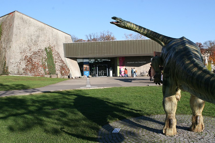The Museum am L&ouml;wentor with a dinosaur statue in the foreground and visitors at the entrance., &copy; SMNS, R. Baumann