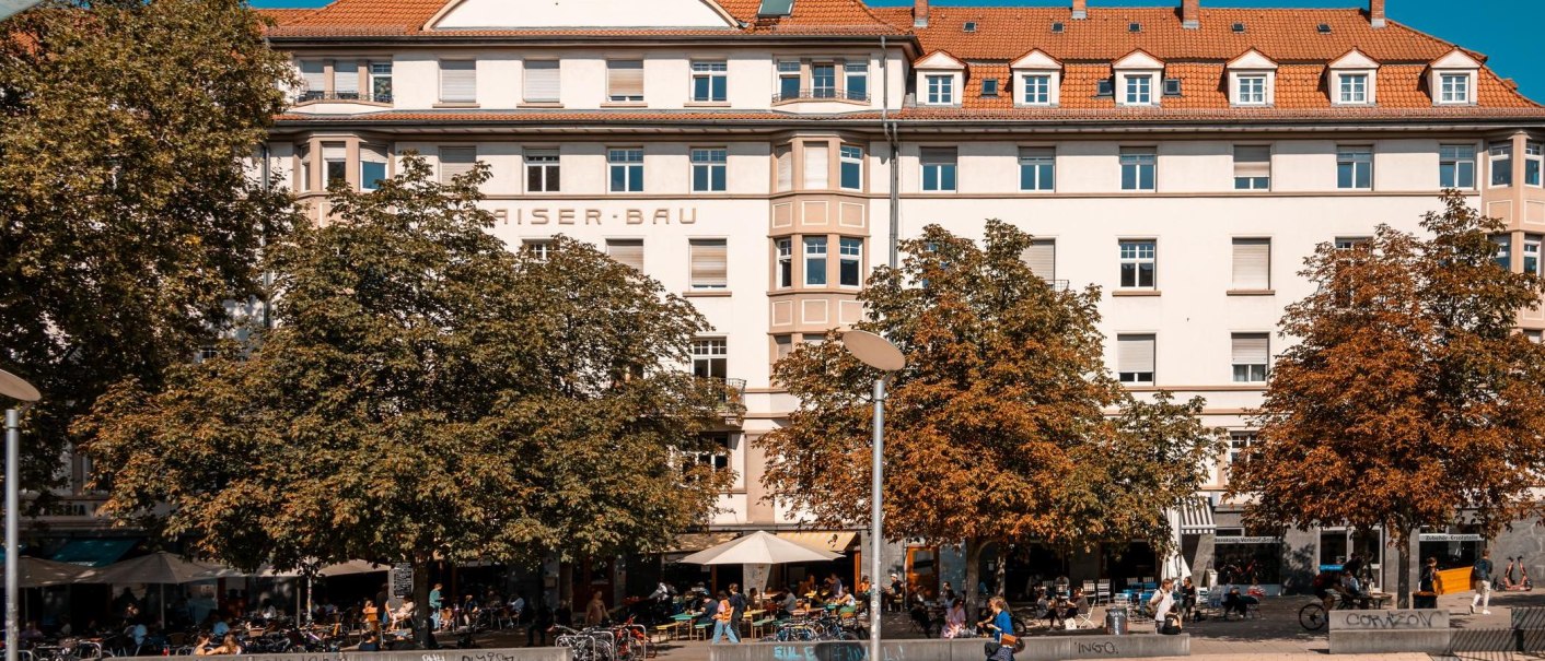 Ein großes Gebäude mit der Aufschrift 'Kaiserbau'. Vor dem Gebäude sind Bäume und Menschen, die in einem Café sitzen. Es ist ein sonniger Tag., © Stuttgart Marketing GmbH, Sarah Schmid