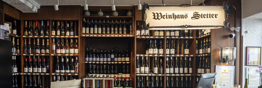 Counter at Weinhaus Stetter with shelves full of wine bottles and a wooden counter. A sign reading 'Weinhaus Stetter' hangs from the ceiling., © SMG Stuttgart Marketing GmbH - Sarah Schmid