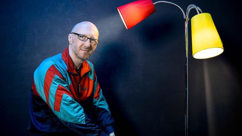 Person in bunter Jacke sitzt neben einer Stehlampe mit roten und gelben Lampenschirmen vor dunklem Hintergrund., © Rosenau Kultur e.V. Person in bunter Jacke sitzt neben einer Stehlampe mit roten und gelben Lampenschirmen vor dunklem Hintergrund., © Rosenau Kultur e.V.