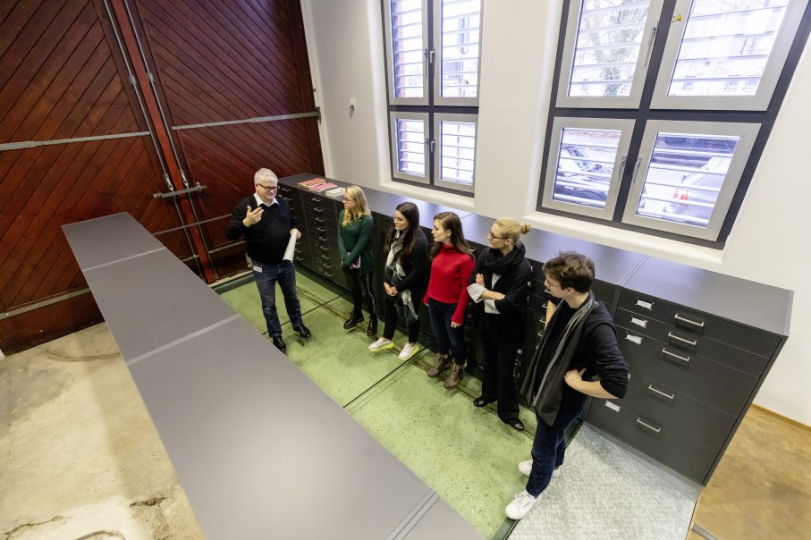 Eine Gruppe von sechs Personen steht in einem Archivraum. Ein Mann erkl&auml;rt etwas, w&auml;hrend die anderen aufmerksam zuh&ouml;ren. Gro&szlig;e Schr&auml;nke und Fenster sind sichtbar., &copy; Stadtarchiv Stuttgart; Foto: arge lola