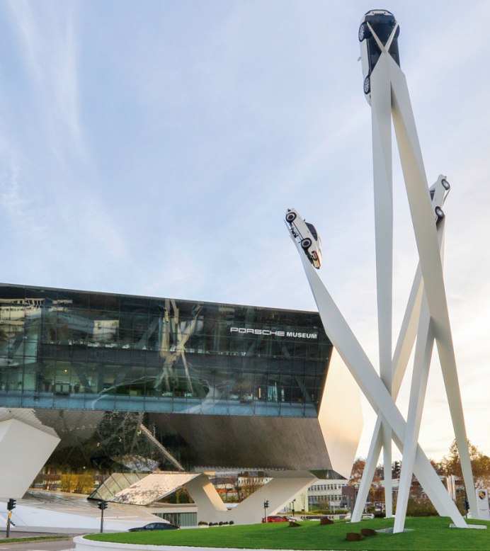 The Porsche Museum with its modern architecture and a sculpture of three cars on high pillars against a blue sky., © Porsche AG, Sabine Braun