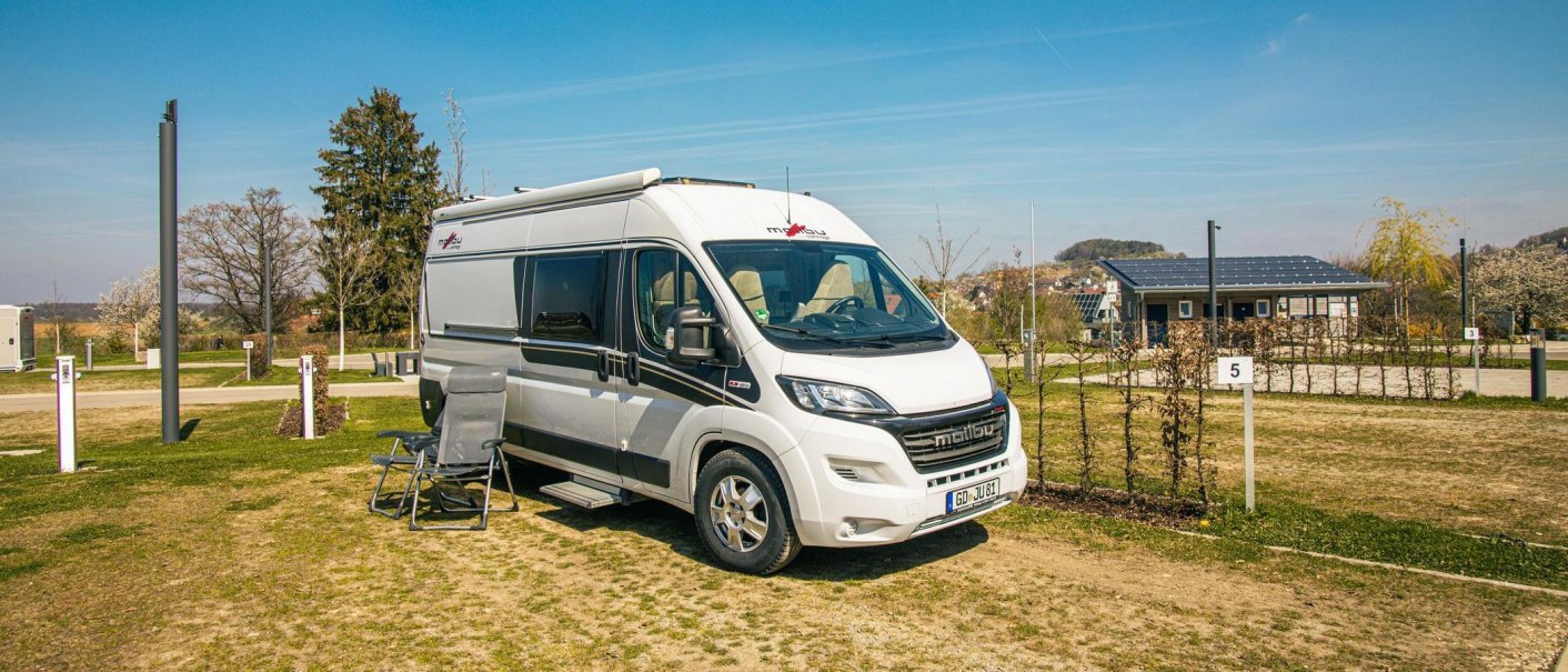 A motorhome is parked on a pitch in Beuren. Chairs and a table are set up next to it. Trees and a building with solar panels can be seen in the background., © Stuttgart-Marketing GmbH, Sarah Schmid
