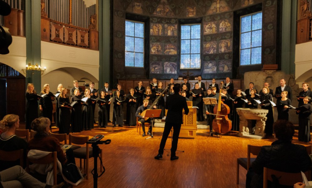 Ein Chor singt in einer Kirche, dirigiert von einer Person. Zuschauer sitzen auf St&uuml;hlen im Vordergrund und lauschen der Darbietung., &copy; 2024 Heiko Tiedmann