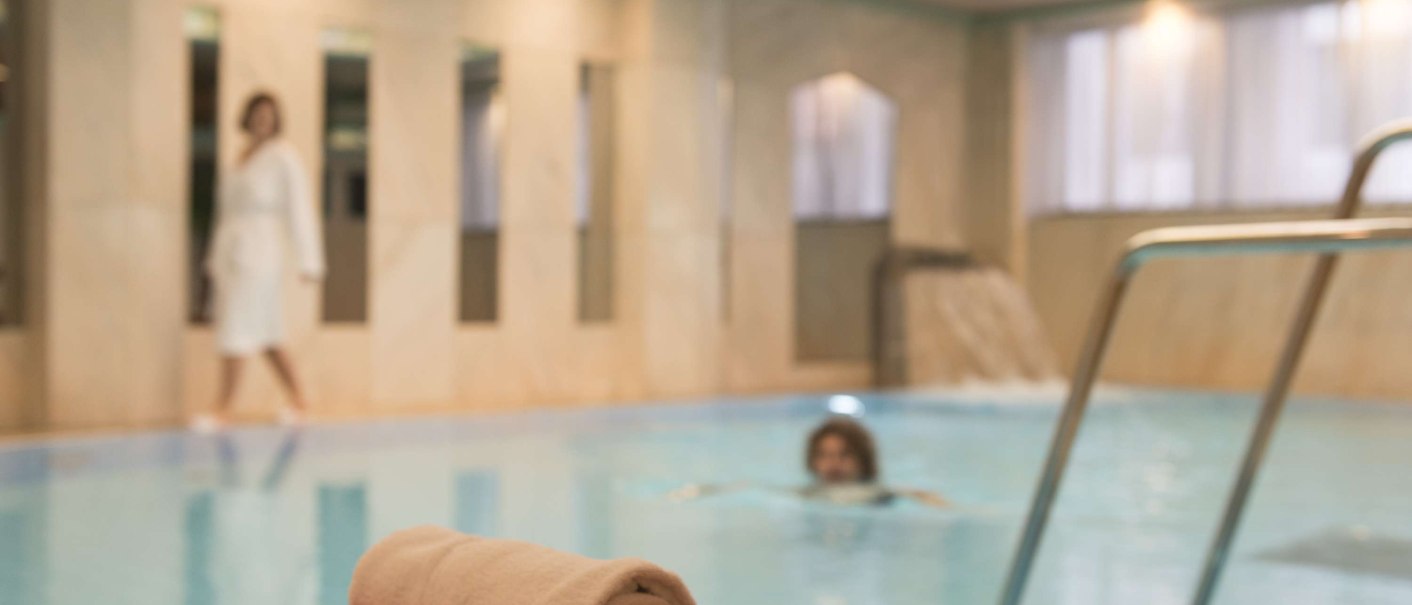An indoor pool with a folded towel on a red cloth in the foreground. In the background a person is swimming, another is walking in a bathrobe., © Maritim Hotelgesellschaft mbH An indoor pool with a folded towel on a red cloth in the foreground. In the background a person is swimming, another is walking in a bathrobe., © Maritim Hotelgesellschaft mbH