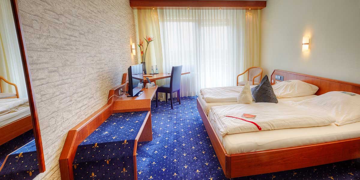 Ein gemütliches Doppelzimmer mit zwei Betten, Schreibtisch, Fernseher und großen Fenstern mit Vorhängen. Teppichboden und helle Wände., © Kemnater Hof GmbH