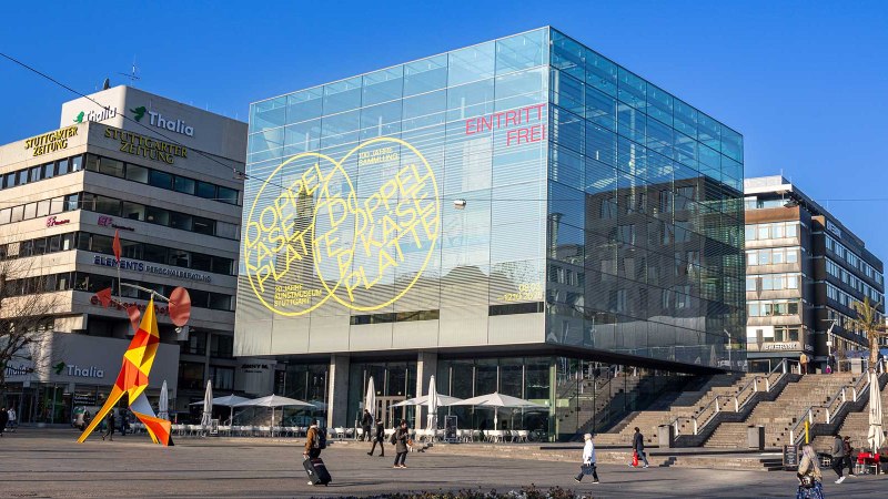 The Stuttgart Art Museum with its striking glass cube on Schlossplatz. A colorful sculpture stands in the foreground, surrounded by modern buildings., © Stuttgart-Marketing GmbH, Sarah Schmid