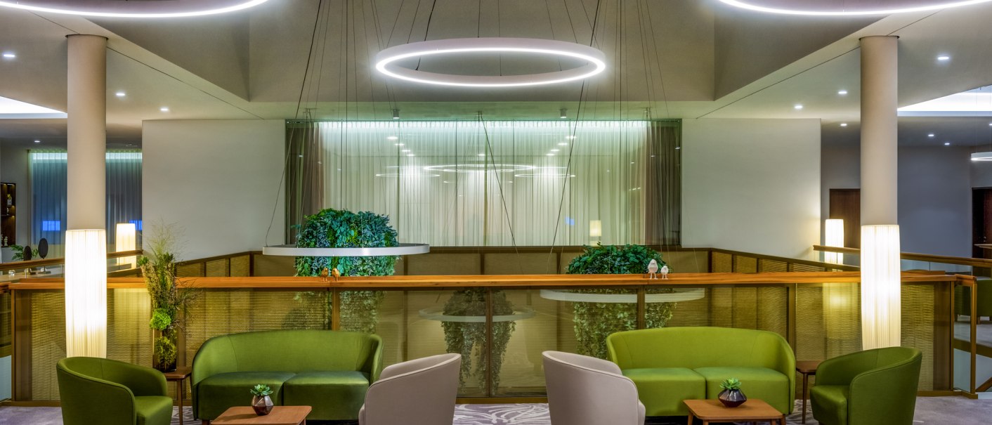 Moderne Lounge mit grünen und beigen Sesseln, runden Tischen und großen, runden Deckenleuchten. Pflanzen und Teppich mit floralem Muster., © Mövenpick Hotel Stuttgart Messe & Congress