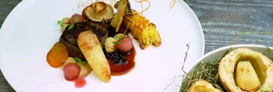 Gourmet dish on a white plate: meat, vegetables, sauce and elaborate garnish. Next to it, a small bowl with pastries and potatoes on a wooden base., © Speisemeisterei
